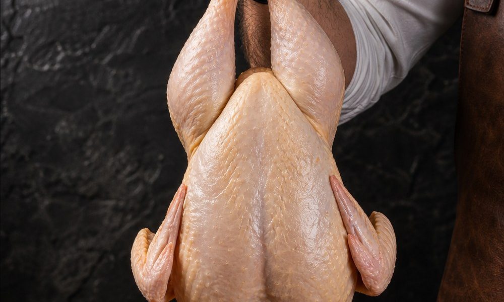 Premium Poultry butcher in apron lifting a whole raw chicken with a black-gloved hand